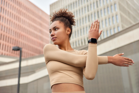 The Benefits of Daily Movement: How Just 30 Minutes Can Transform Your Health 42 curly haired young woman stretches arms warms up before workout dressed cropped top smartwatch poses outdoors